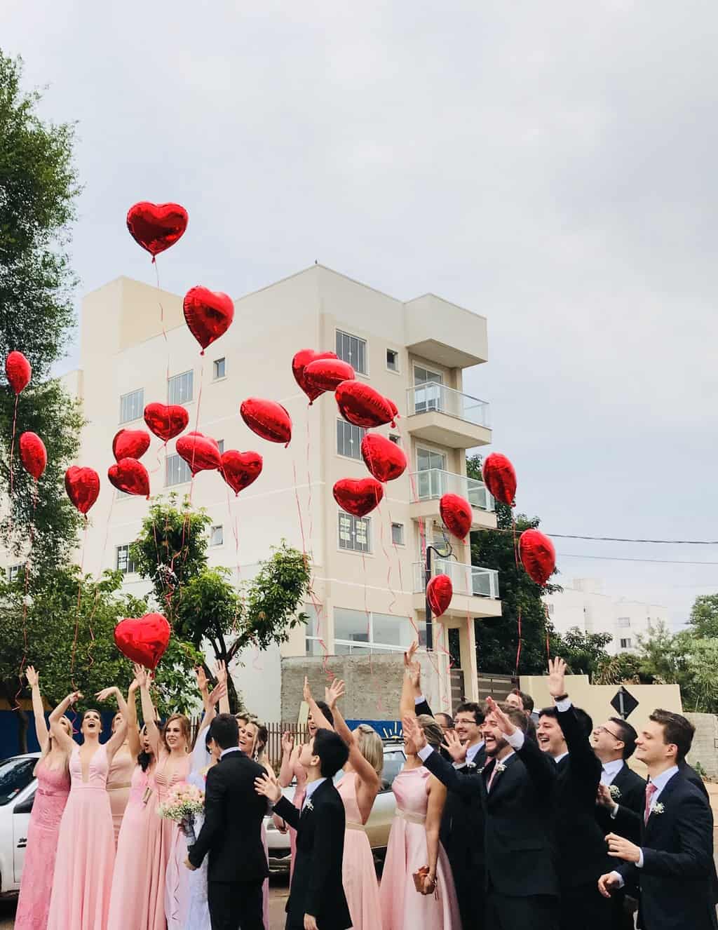 Women Wearing Pink Dresses and Men Wearing Black Suit Jacket and Pants Raising Hands With Red Heart Balloons
