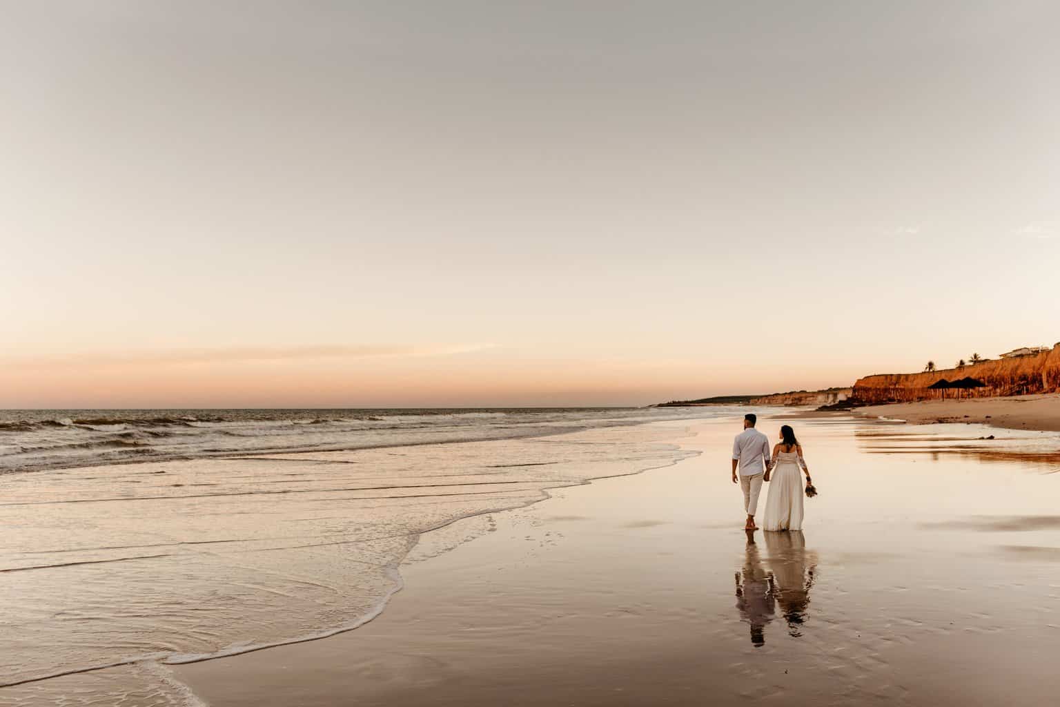 Free Back view of barefoot groom holding hand of bride in white dress and walking together on sandy coast Stock Photo