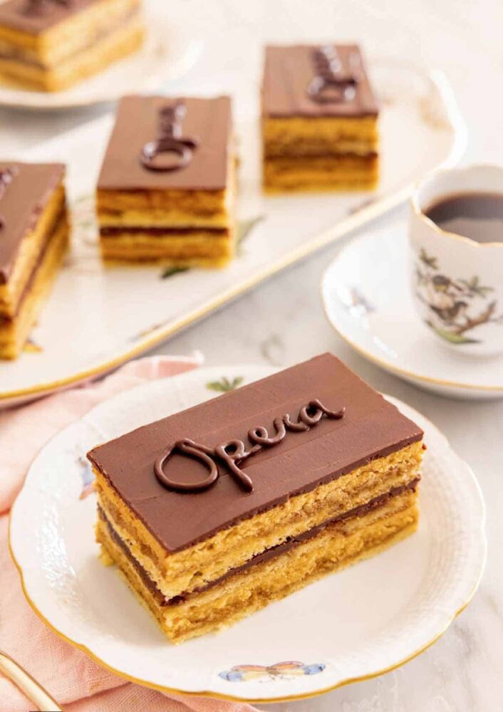 A serving of opera cake on a plate by a cup of coffee with more cakes in the background.