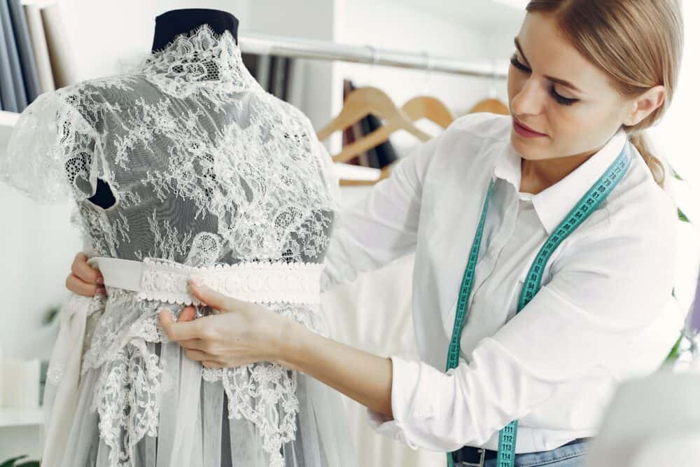 Free Talented female tailor wearing white shirt and jeans working with mannequin in modern atelier or wedding boutique while attaching belt on lace dress Stock Photo