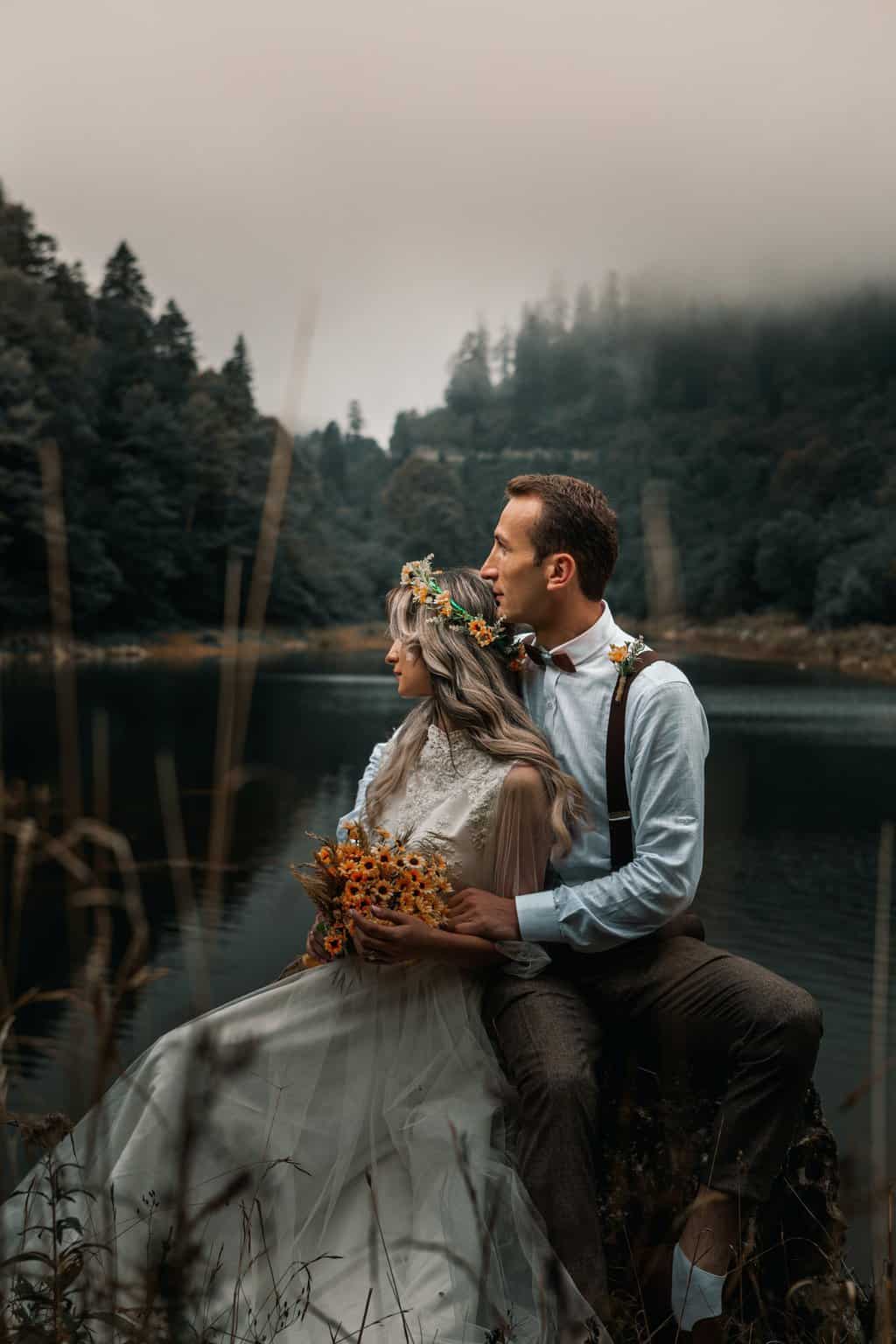 Free Young groom gently embracing bride with flower wreath and bouquet while sitting on river shore against woods and looking away Stock Photo